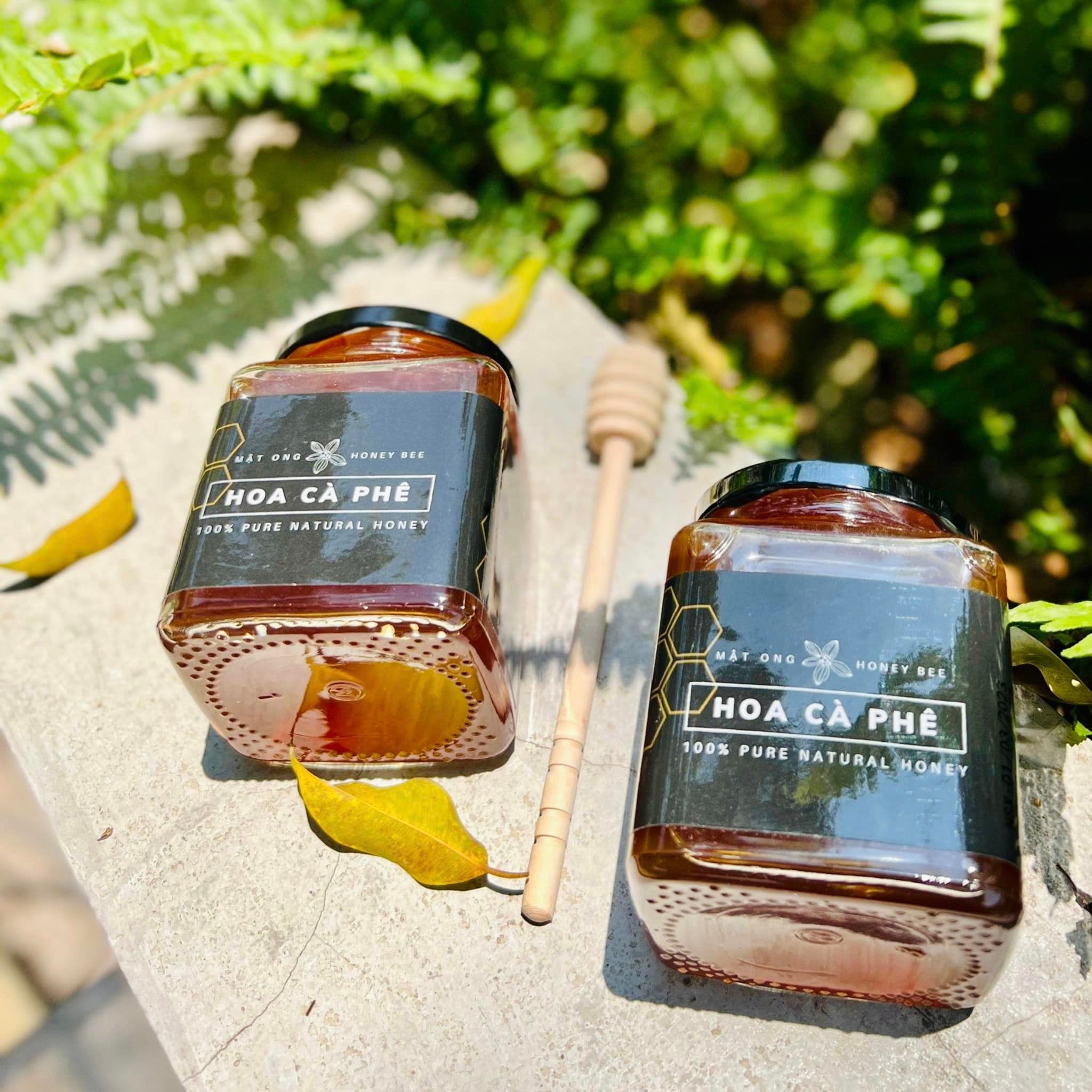 Cận cảnh giọt mật ong hoa cà phê nguyên chất óng vàng trên nền hoa cà phê trắng tinh khôi, nổi bật sự tinh khiết và tự nhiên của sản phẩm.